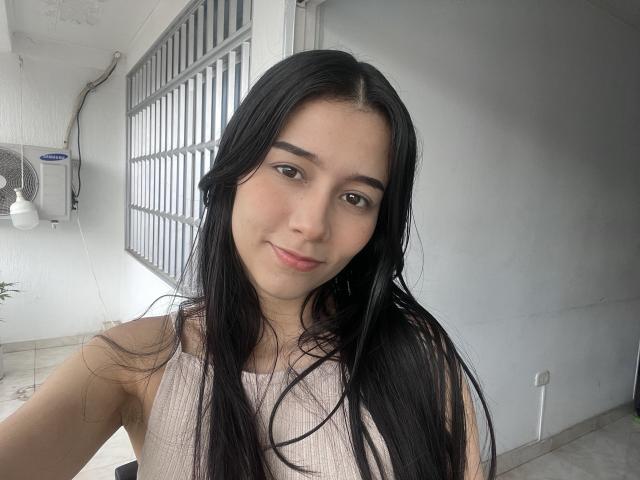 DeisyWest with long black hair, brown eyes, and a slight smile, wearing a pink ribbed tank top, showing her small tits, in a gray-walled room with a barred window.