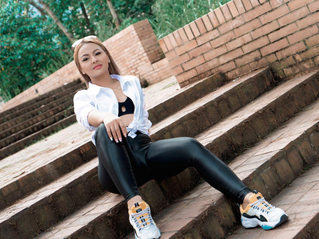 AmeliaMerie, con sus glúteos sobre unos escalones de ladrillo, vistiendo una camisa blanca, una camiseta negra, pantalones de cuero negro y zapatillas blancas con detalles amarillos, mirando a la cámara..