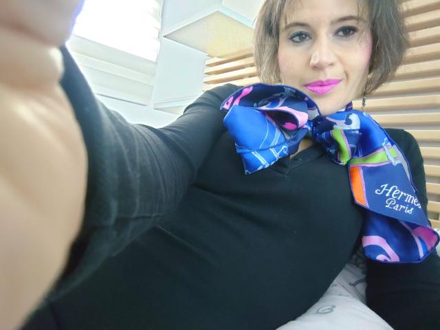 SoniaHayat with pink lipstick, wearing a black top and colorful blue scarf, taking a selfie, showing her chest, smiling, in a bright room with wooden slats.
