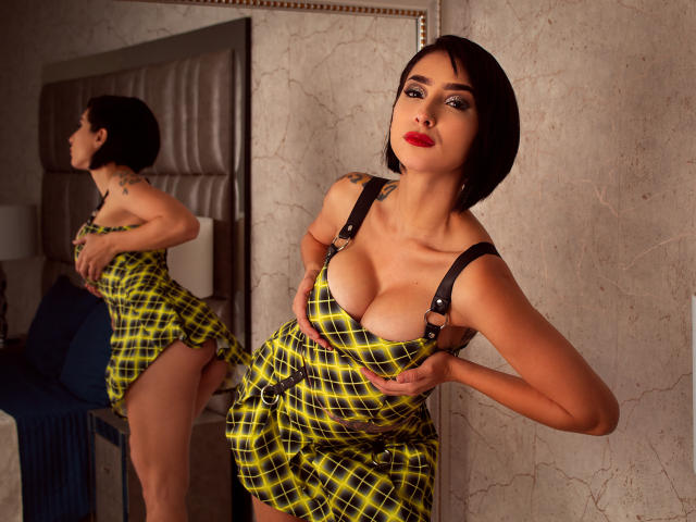 LolaGinger, com cabelo preto curto, batom vermelho e um vestido xadrez amarelo, está segurando seus seios grandes, mostrando seu traseiro em um reflexo no espelho. Ela está em pé em um quarto..