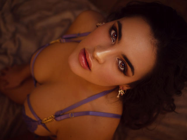 Celeste Belucci, com olhos castanhos, vestindo lingerie roxa, tem seios grandes e está olhando para cima com lábios rosados, cabelos escuros e ondulados, e brincos de ouro, em uma foto íntima com iluminação quente..