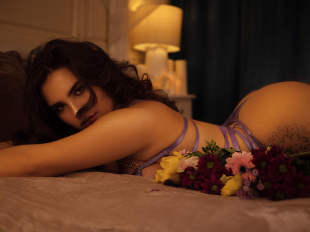 Celeste Belucci, avec ses cheveux noirs bouclés, est allongée sur un lit en lingerie violette, regardant de manière séduisante l'appareil photo, avec un bouquet de fleurs devant elle, dans une pièce faiblement éclairée..