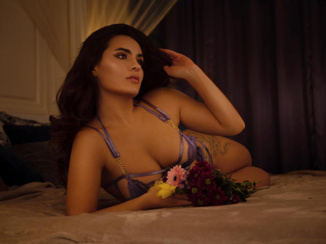 CelesteBelucci with dark hair, wearing purple lace bra, lying on bed, holding flowers, tattoo on side, looking to the side, hand in hair, soft brown bed, dark curtains.