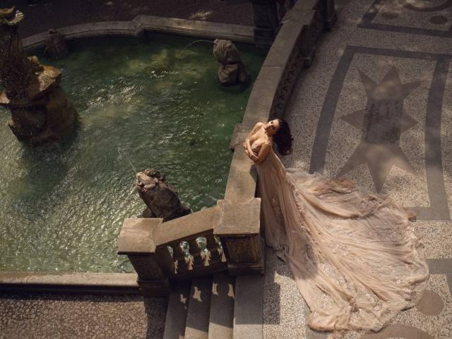 Celeste Belucci mit großen Brüsten in einem fließenden, beige farbenen, spitzenkleid, die sich an ein steinernes Geländer lehnt, neben einem grünen, mit Wasser gefüllten Brunnen mit Steinfiguren. Darüber, geflecktes Sonnenlicht..