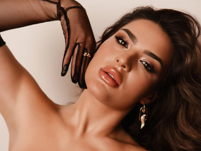 CelesteBelucci with glossy brown lips, dark eyeliner, and shiny skin, wearing black mesh gloves, gold ring, and dangling earrings, looking seductive with her hand near her face.