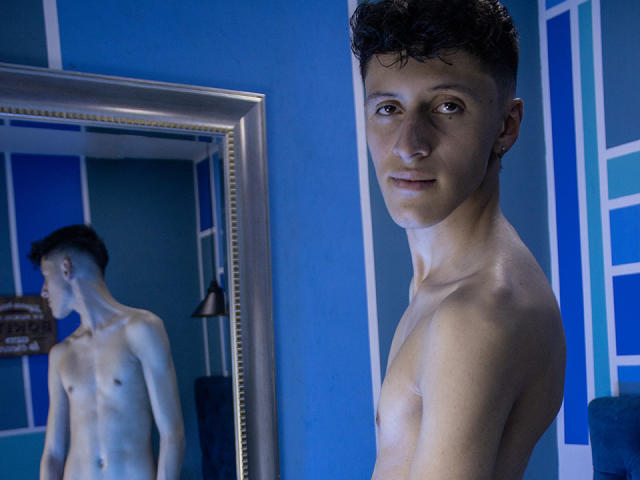 Carlos Rubiio, sem camisa, com cabelo curto, cacheado e escuro, em pé em um quarto azul, refletido em um espelho, mostrando um corpo magro, tonificado e pálido, com nádegas pequenas e seios pequenos, olhando para a câmera..