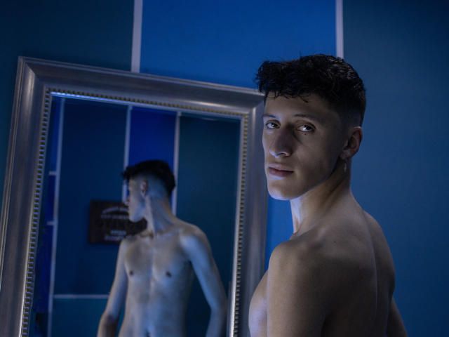 Carlos Rubiio, senza camicia, in piedi di fronte a uno specchio, il riflesso mostra un ragazzo bianco, magro e in forma, con capelli neri ricci, occhi azzurri, un leggero sorriso, una stanza blu, lo specchio ha una cornice argentata..