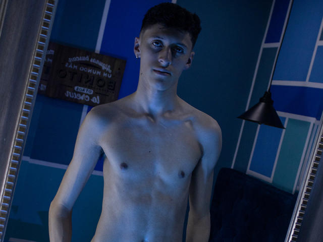 CarlosRubiio, shirtless, blue-tinted, has a slim, toned body with small tits, short dark hair, and a slight smile, standing in front of a mirror with a black lamp.