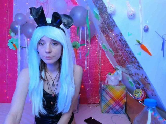 MissAstravert, con cabello blanco, orejas de conejo negras y una blusa de cuero negro, está sentada en una habitación iluminada con luz azul, con globos, decoraciones y un regalo. Sus pechos son pequeños y su vulva está oculta..
