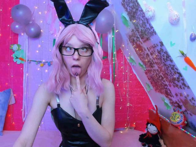 MissAstravert, avec ses cheveux roses, ses oreilles de lapin noires, ses lunettes et sa robe en cuir noir, fait un geste de silence en posant son doigt sur ses lèvres, tout en exhibant sa petite poitrine dans une pièce colorée et festive..
