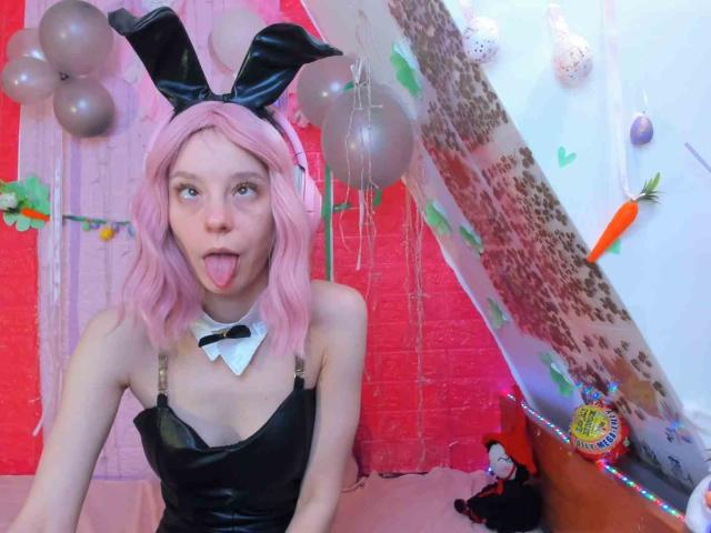 MissAstravert, com cabelo rosa, orelhas de coelho de couro preto e vestido preto, de língua para fora, usando uma gola branca e laço preto, em um cosplay divertido e sexy..