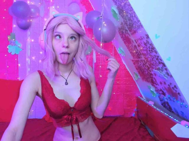 MissAstravert with pink hair, cat ears, and headphones, sticking out her tongue, wearing a red lace bra, holding her hair, and sitting on a red bed.