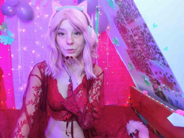 MissAstravert con i capelli rosa, orecchie da gatto, top di pizzo rosso che mostra il seno, con la mano sul mento, circondata da luci rosa e viola, con le cuffie, dall'aspetto carino..