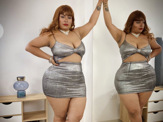 SammyHank has big boobs, a curvy ass, and a small cutout in her silver, shiny, mini-dress. She's standing with one arm raised, reflecting in a mirror.