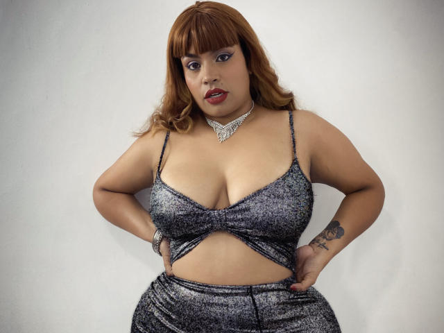 SammyHank, curvy woman with brown hair, wearing a sparkly grey crop top and matching pants, showing her big tits, choker necklace, tattoo on right hand, hands on hips.