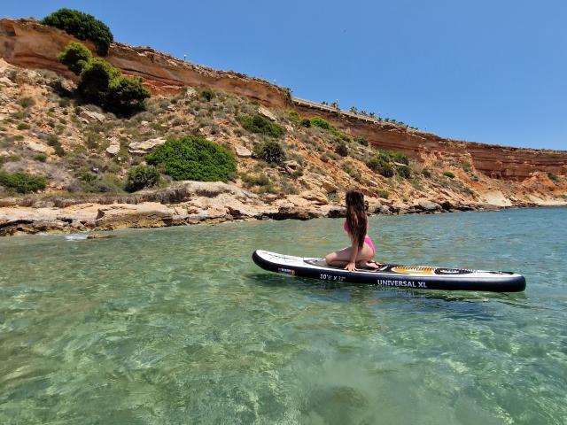 ValeriMair with long brown hair, wearing pink bikini bottoms, sitting on a black and white stand-up paddleboard in clear, shallow water, facing rocky shoreline under bright blue sky.