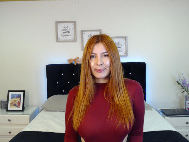 AnnyCraffort with long red hair, medium tits, wearing a red long-sleeve shirt, smiling at the camera, sitting on a bed with a black headboard.