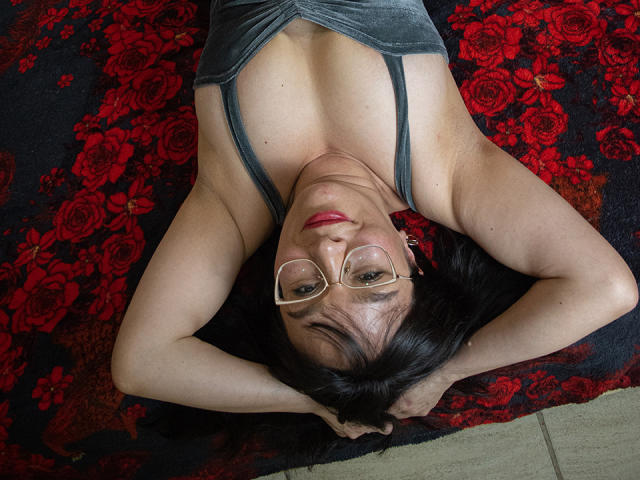 LexiLuna, with glasses and red lipstick, lies on a red floral rug, arms above her head, wearing a grey tank top, showing her tits and looking up.