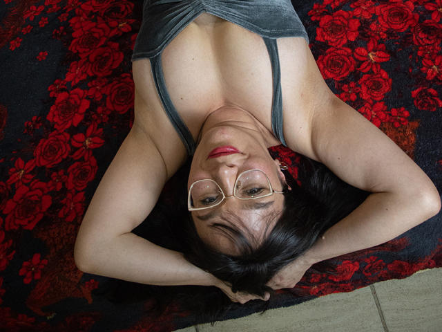 LexiLuna ligt op haar rug met haar armen boven haar hoofd, ze draagt een bril en een grijze top, waarbij haar borsten zichtbaar zijn en ze rode lippenstift draagt, op een rood-zwarte bloemenkleed..