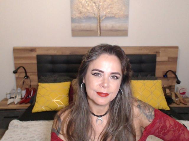 EvaLuv has long brown hair, red lipstick, and red lace top, with tattoos on her right arm, sitting on a bed with yellow pillows and wooden headboard.