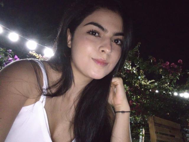 Marilyn Woods met lang zwart haar, een witte tanktop, kleine borsten, een schattig gezicht, een lichte glimlach, een zwarte armband, 's nachts, met lichtjes en roze bloemen op de achtergrond..