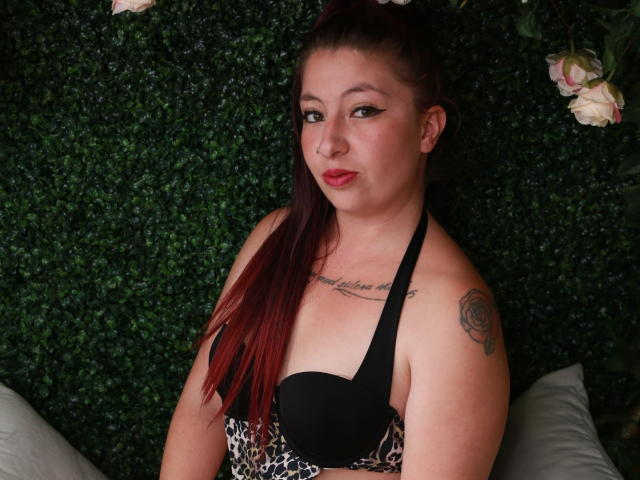 Nicoleks com cabelo ruivo, sutiã preto e com estampa de leopardo, tatuagem no ombro direito, olhando para a câmera, parede com folhagem verde, lábios levemente entreabertos, maquiagem discreta, expressão confiante..
