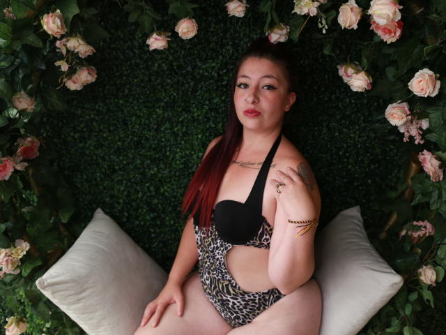 Nicoleks mit roten Haaren, schwarzem BH und einem einteiligen Badeanzug mit Leopardenmuster, sitzt auf weißen Kissen, mit Blumen und Grünpflanzen im Hintergrund, hat ein Tattoo an der rechten Schulter, ein Armband am linken Handgelenk und eine Hand in der Nähe des Gesichts..