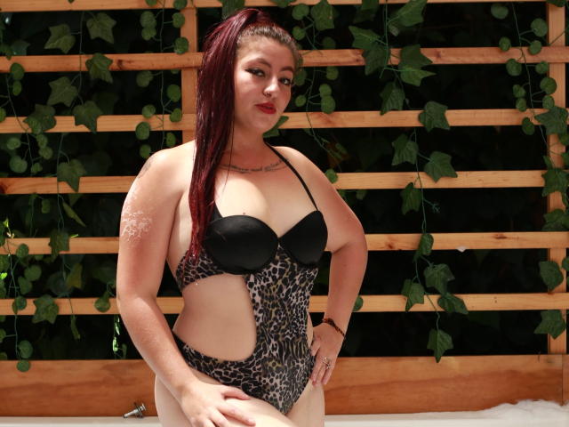 Nicoleks with red hair in a leopard print one-piece, black bra, showing her tits and ass, standing in a hot tub, looking at the camera, hands on hips.