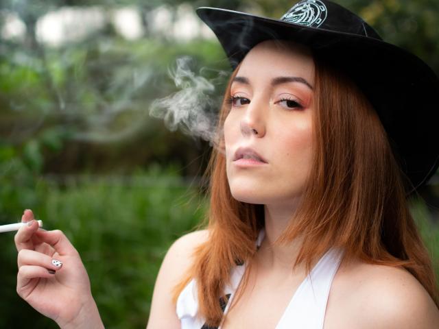 MollyMarmalade with red hair, wearing a black hat, smoking a cigarette, holding it near her nose, white halter top, green background, slight smoke, looking bored.