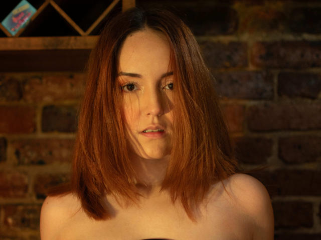 MollyMarmalade with shoulderless top, brown hair, slightly wavy, small nose, subtle makeup, looking slightly to the side, soft light, brick wall background, slightly parted lips.