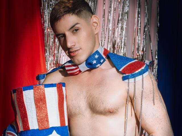 JonathanJeremiah, sin camisa y con adornos de la bandera estadounidense, sosteniendo su entrepierna cubierta con la bandera, cabello castaño corto, ligera sonrisa, cadenas plateadas, fondo rojo y cortina plateada brillante..