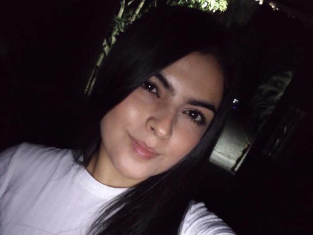 MarilynWoods ha lunghi capelli neri, occhi marroni e un leggero sorriso. Indossa una camicia bianca, con un trucco leggero che mette in risalto il suo seno carino e piccolo. Sfondo scuro..