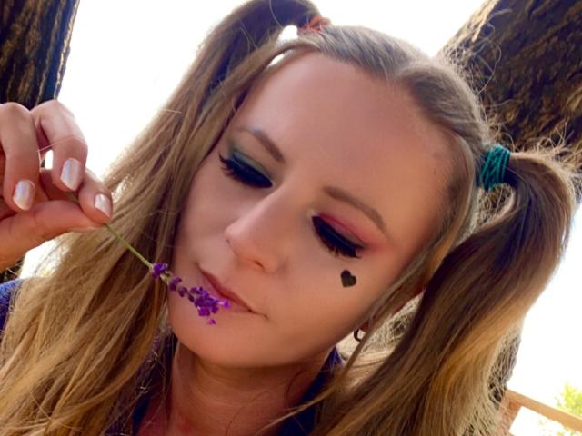 LaMadrina with puffy blue and pink eyeshadow, long blonde pigtails, white nail polish, small black heart tattoo, holding purple lavender, slightly pouty lips, wooden background.