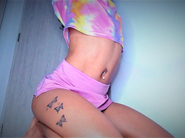 KarenSofia69 in pink tie-dye top, pink shorts, showing her toned ass, butterfly tattoos on right thigh, belly button piercing, light skin, slightly bent forward.