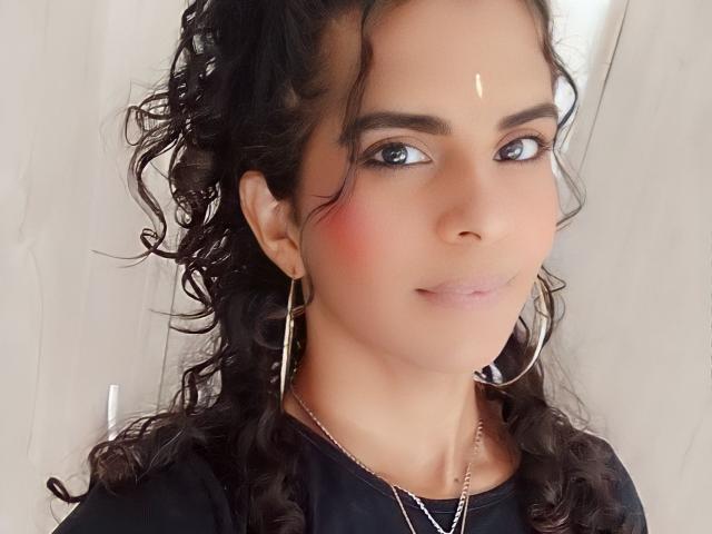 KarenSofia69 tiene el cabello negro y rizado, ojos marrones y una sonrisa sutil. Lleva aretes de aro dorados y una blusa negra con una pequeña marca blanca en la frente..