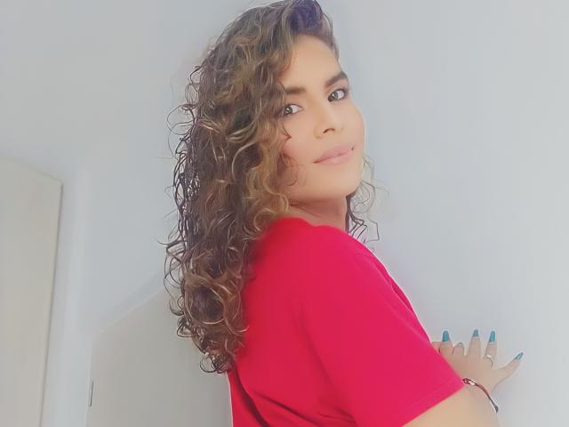 KarenSofia69 with curly brown hair, wearing a red shirt, looking back with a slight smile, blue nails, white wall, and a subtle ring on her right hand.