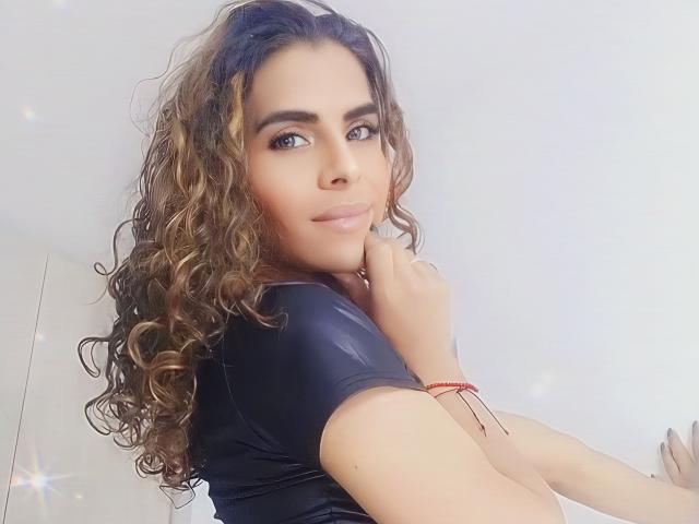 KarenSofia69, con cabello castaño rizado, vistiendo una camisa negra, mirando por encima del hombro, sonriendo, sosteniendo su barbilla, con una pulsera roja en la muñeca, piel clara, maquillaje sutil, labios rosados suaves..