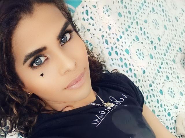 KarenSofia69 hat lockiges, braunes Haar, blaue Augen und ein kleines, schwarzes Herz auf ihrer Wange. Sie trägt ein schwarzes Hemd mit einer Halskette und liegt lächelnd da..