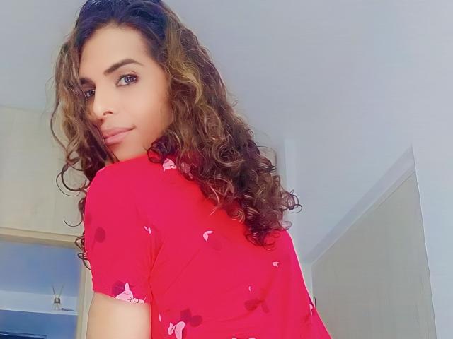 KarenSofia69, con cabello castaño rizado, vistiendo una camisa roja con estampados de mariposas rosas, mirando por encima del hombro, sonriendo ligeramente, en un ambiente interior luminoso..