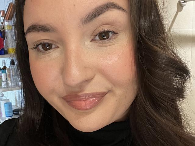 Het gezicht van ParadisCamille, met bruine ogen, donkerbruin golvend haar en glanzende roze lippen, met een lichte glimlach en een kleine puist op haar rechterwang..