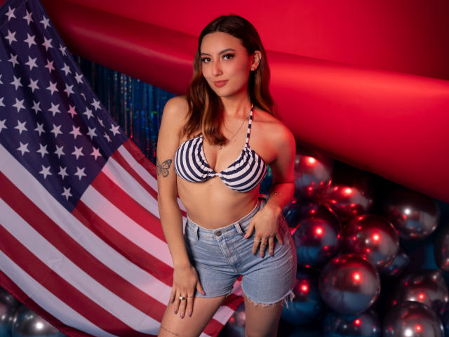 CarlaFranco with striped bikini top, showing her tits, in denim shorts, standing in front of a large American flag, surrounded by shiny silver balloons, under red lighting.
