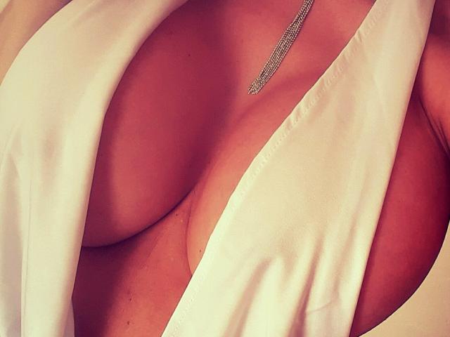 I seni grandi e rotondi di LaParisienne sono appena coperti da un abito bianco e fluente, con una collana d'argento che pende appena sopra il suo petto. L'immagine si concentra sul suo décolleté..