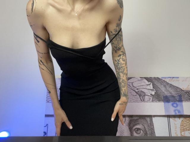 Tsukerberg, avec des tatouages sur les bras, portant une robe noire moulante sans bretelles, les mains sur les hanches, avec un décolleté visible, la clavicule et un collier. Peau transparente, silhouette mince, légèrement penchée en avant..