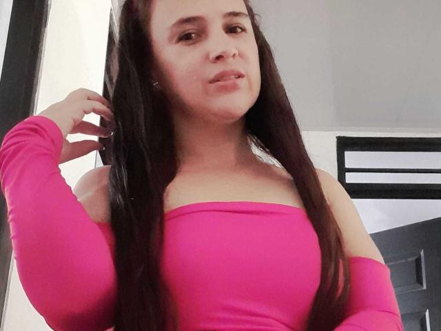 Brihana Charlot, avec ses longs cheveux foncés, son haut rose à une épaule, et son petit anneau de nez, souriant légèrement, la main droite dans les cheveux, montrant ses seins et une partie de ses fesses..