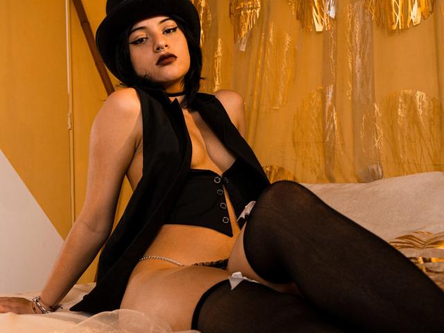 DaliaBelle, avec sa coupe de cheveux courte noire et son chapeau, porte un haut noir, un corset et des bas hauts, exposant ses seins et montrant sa vulve avec une chaîne, assise sur un lit..