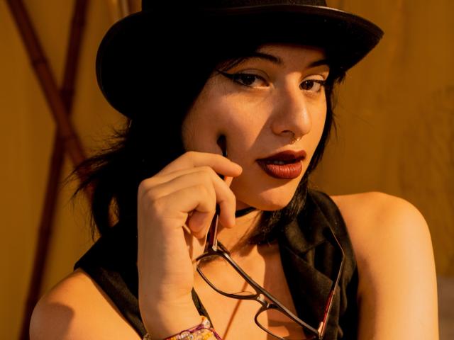 DaliaBelle mit schwarzem Hut, dunklem Lippenstift, die eine Brille hält, trägt ein schwarzes Top ohne Ärmel, hat eine Nasenring und ein rosa Armband und blickt leicht zur Seite, in warmem Licht..