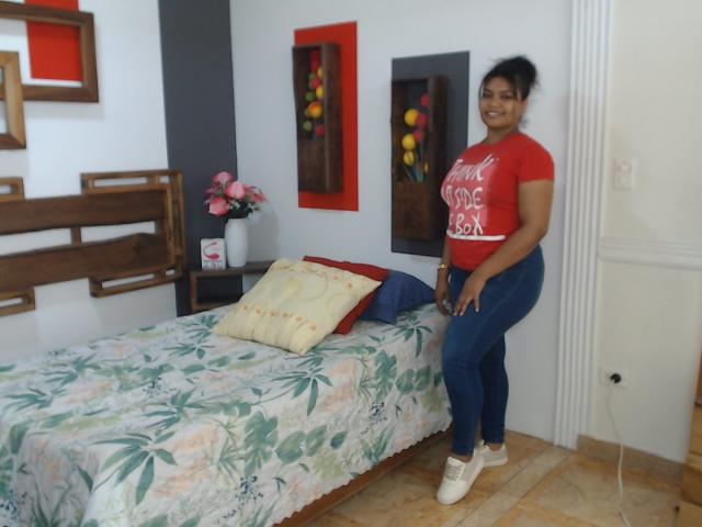CataleyaX69 with dark skin, black hair in a bun, red shirt, blue jeans, and white sneakers, standing next to a floral-patterned bed, smiling. She has a medium ass.