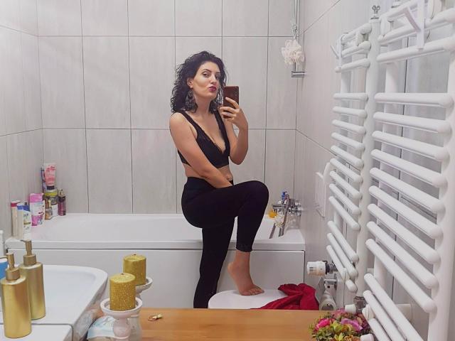 AddictedToUTS, con cabello negro rizado, vestida con un sostén y pantalones negros, sentada en la bañera, tomando una selfie en el espejo, mostrando sus pechos, sosteniendo un teléfono, con un radiador blanco a la derecha..