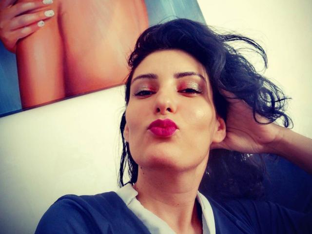 AddictedToUTS a les cheveux noirs et ondulés, du rouge à lèvres, et un fard à paupières brillant, elle fait la moue en se tenant les cheveux, et porte une chemise bleue. L'arrière-plan montre un gros plan d'une fesse bronzée et nue..
