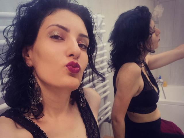 AddictedToUTS with black wavy hair, wearing a black lace bra, pouty red lips, and dangling earrings, standing in front of a mirror, showing her ass in reflection.